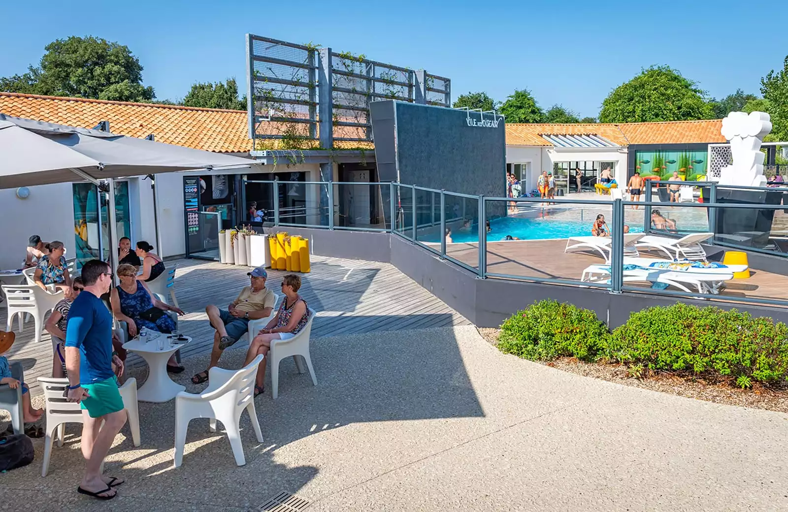 des vacanciers sont dans un espace convivial sous des parasol vu sur la piscine extérieure du camping